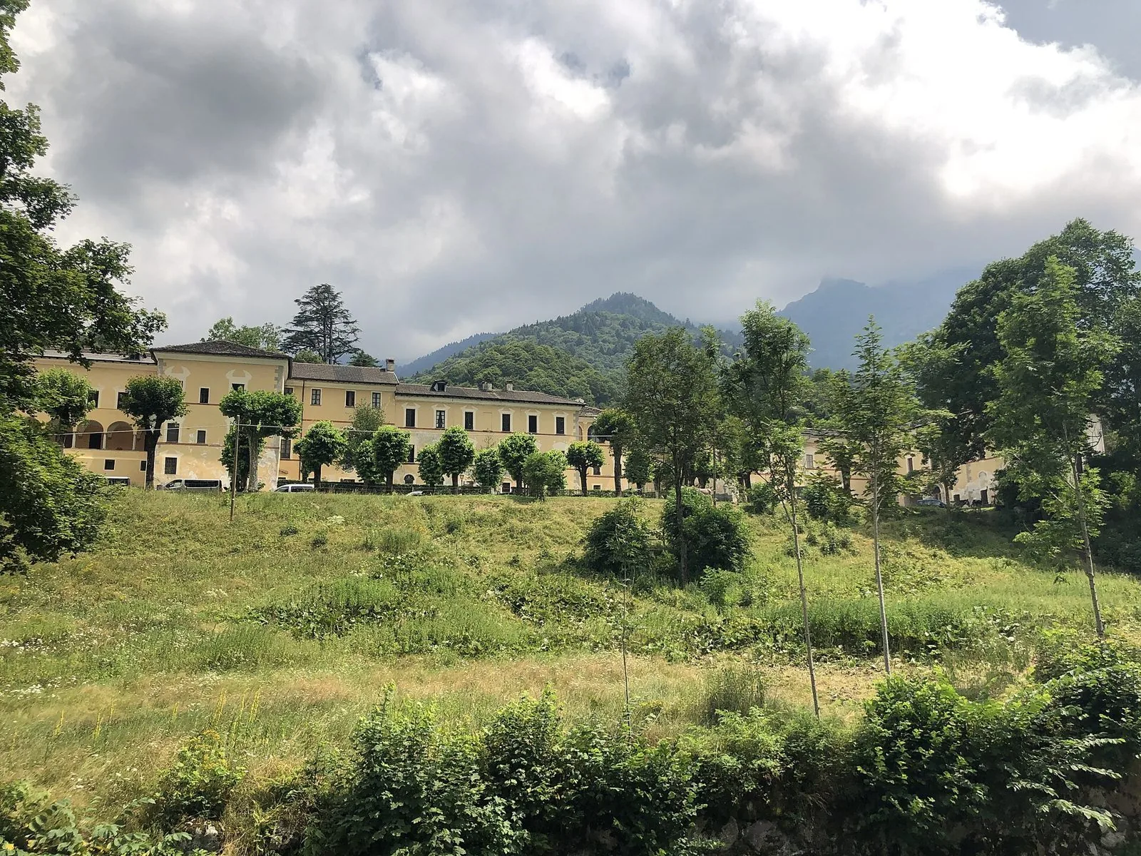 La lunga facciata gialla della Certosa di Pesio tra gli alberi, con le montagne boscose della Valle Pesio sullo sfondo