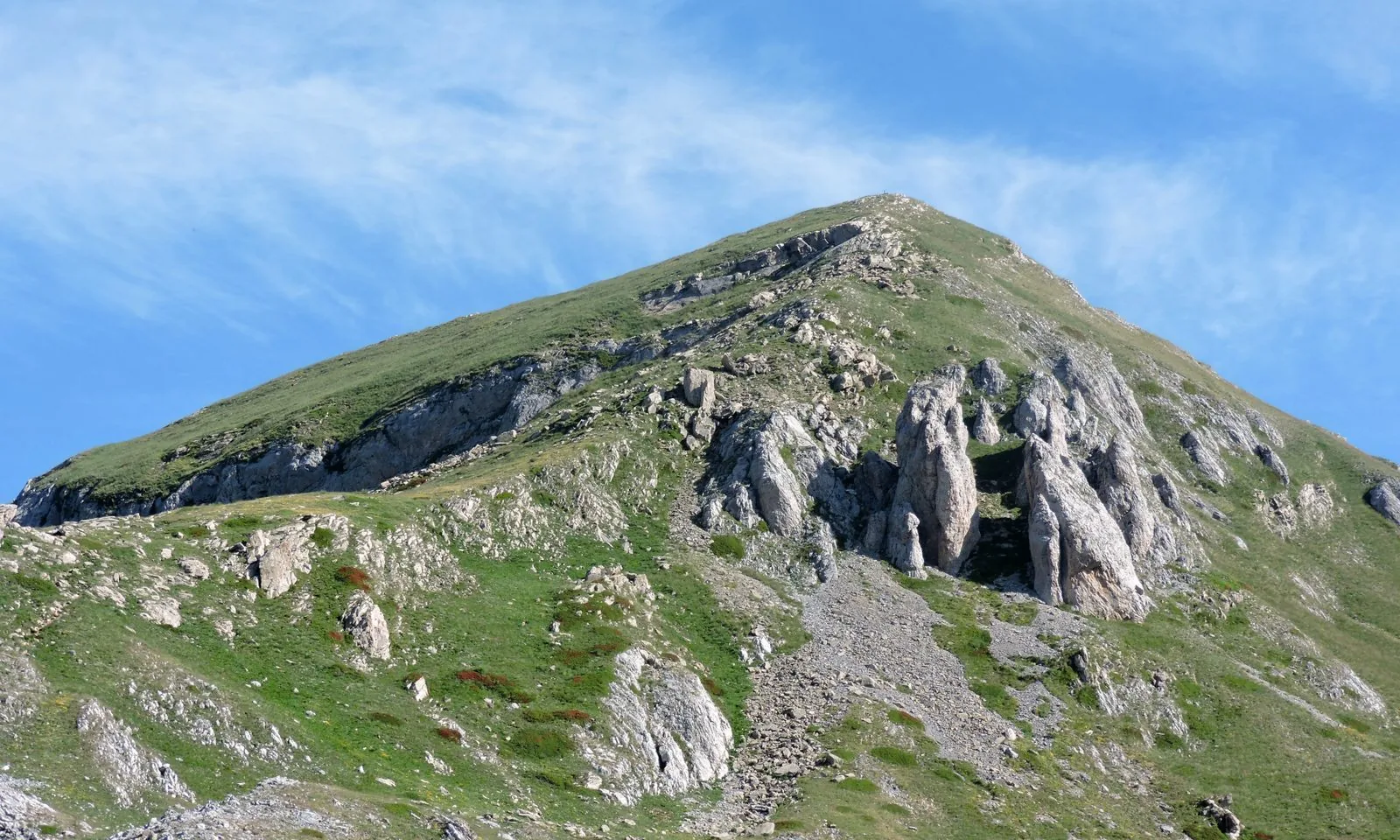 Il versante erboso e roccioso del Monte Mondolè in estate, con guglie calcaree sotto la vetta e cielo azzurro